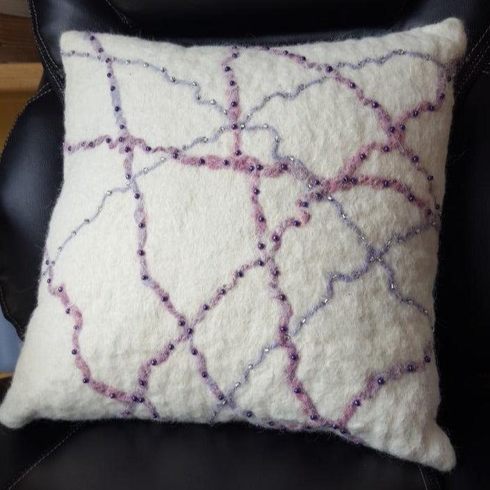 A white felted pillow with random pruple and pink yarns  and glass beads crisscrossing it 